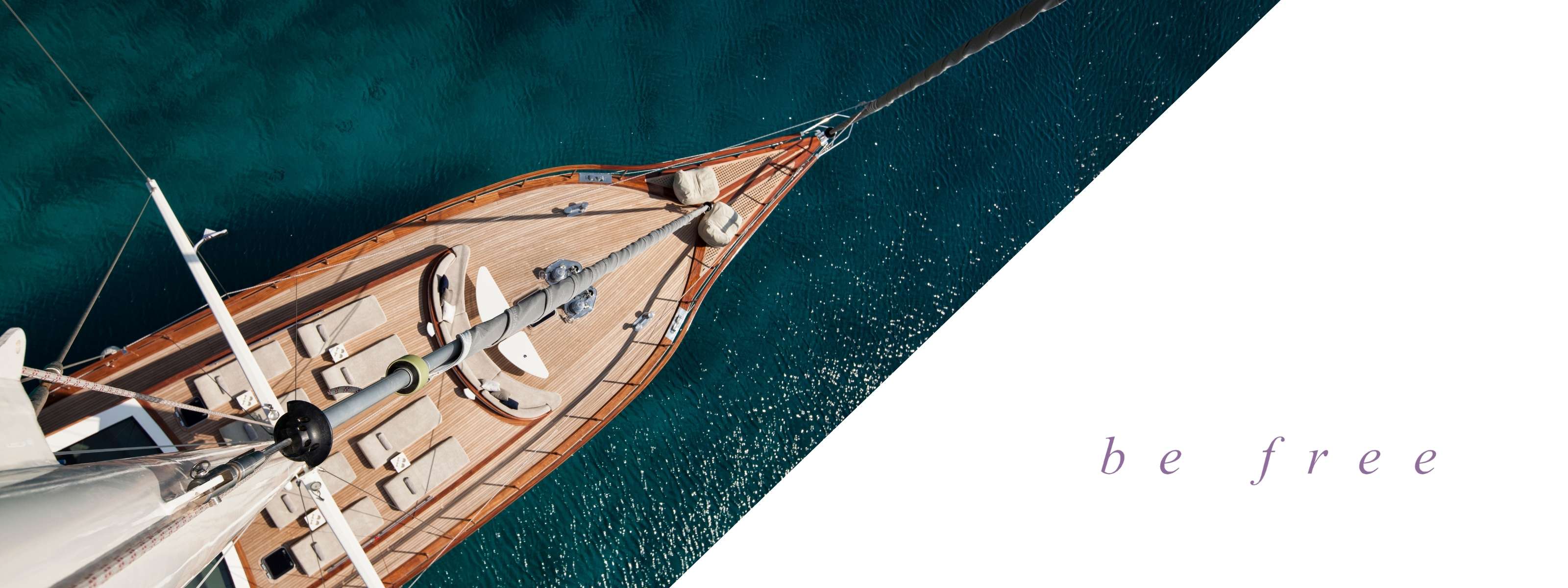 Top-down view of a luxurious wooden sailboat floating on deep turquoise waters. The deck is adorned with plush lounge cushions, evoking relaxation and freedom. The phrase be free in soft lavender text sits elegantly on a white background, enhancing the sense of serenity and escape.