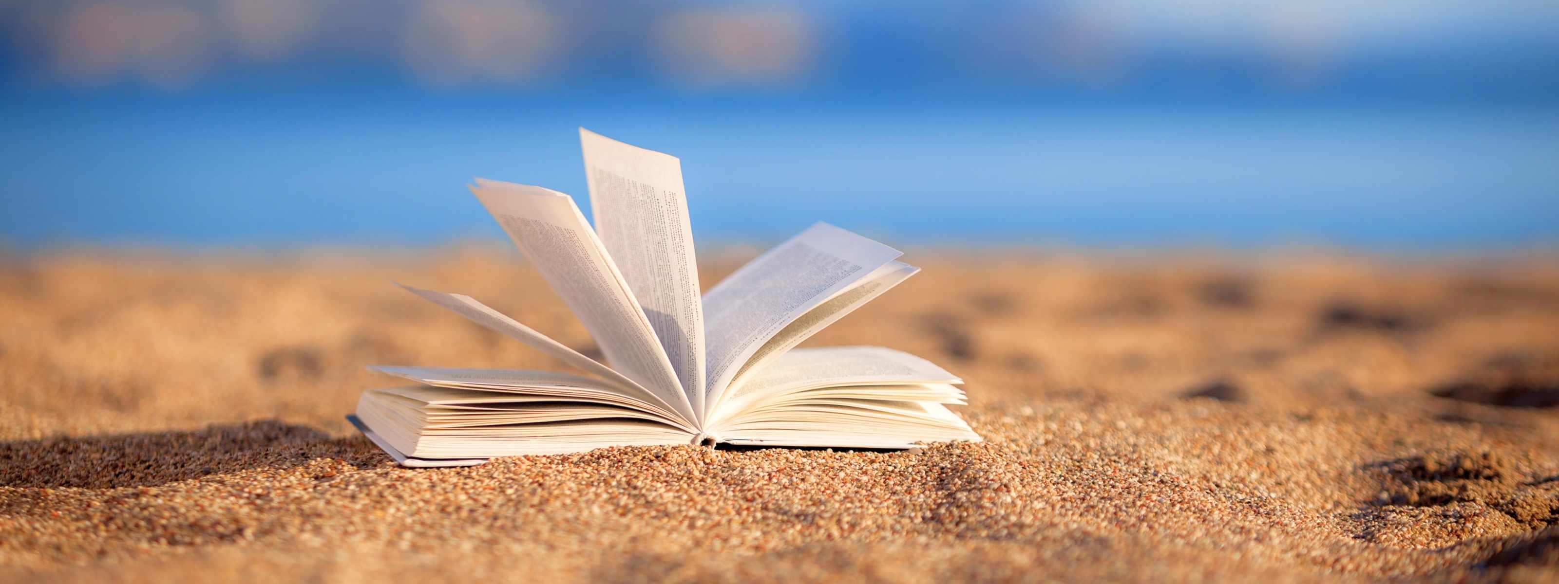 An open book rests gently on golden sand, its pages caught in a soft breeze. The blurred background reveals the tranquil blue of the ocean, blending seamlessly with the sky. The scene evokes a sense of peaceful escape, inviting moments of reflection, relaxation, and the joy of losing oneself in a story by the sea.