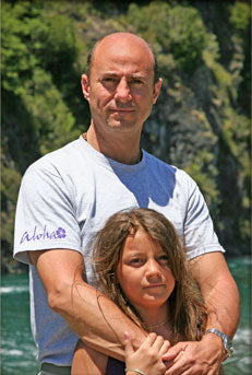 The founder of Aloha standing outdoors in a natural setting, wearing a light-colored shirt with the Aloha logo on the sleeve, holding his daughter in a warm embrace, with lush greenery and cliffs in the background.